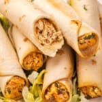 Stack of baked chicken taquitos on a bed of shredded lettuce, one cut in half showing the creamy chicken filling