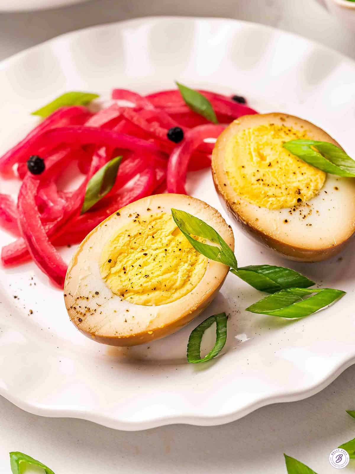 A plate of sliced soy sauce eggs, garnished with scallions and pepper, next to pickled red onions