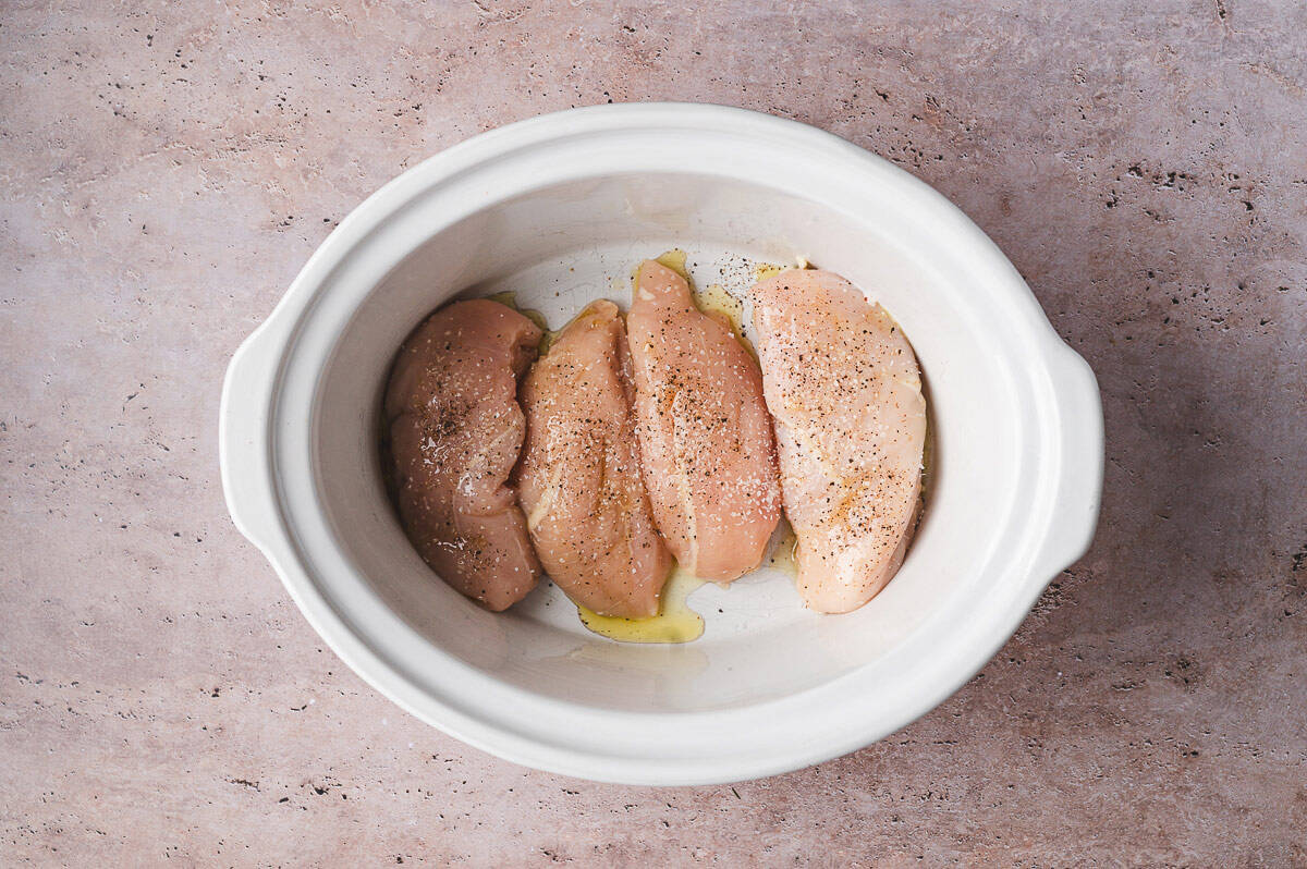 Seasoned raw chicken breasts drizzled with olive oil placed in a slow cooker before adding the remaining ingredients.