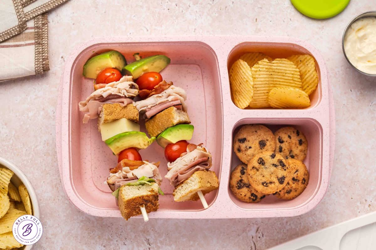 Club sandwich on a stick made with toasted white bread, iceberg lettuce, ham, turkey, bacon, tomatoes, avocado, white cheddar, in a lunch box