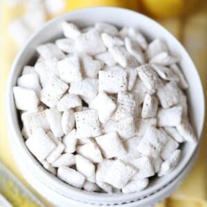 overhead picture of lemon muddy buddies in white bowl with whole lemons and zester on yellow napkin