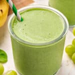 Close up of a green smoothie in a glass jar with a green and white striped straw, surrounded by grapes and a banana