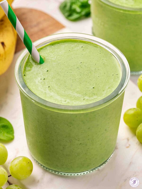 Close up of a green smoothie in a glass jar with a green and white striped straw, surrounded by grapes and a banana