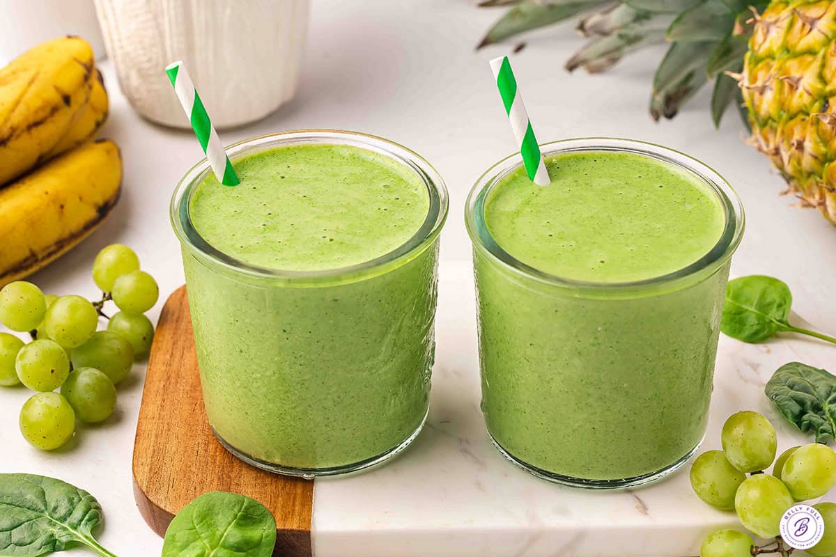 Two green smoothies in glasses with striped straws on a wood and marble board, surrounded by grapes, bananas, and a pineapple