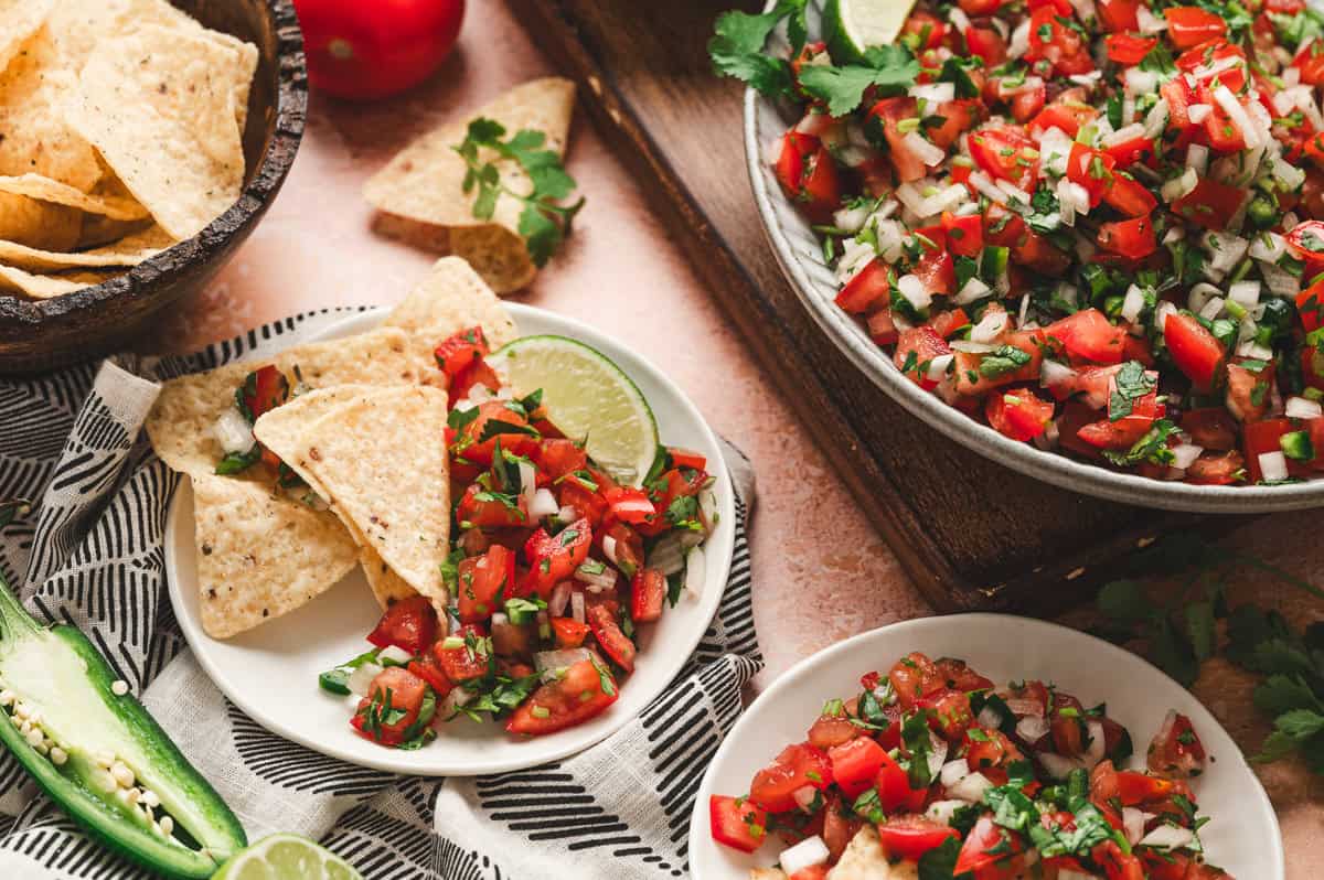 Pico de gallo served with tortilla chips on a plate alongside a bowl of salsa and fresh ingredients