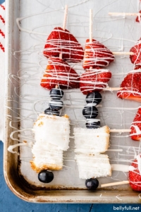 overhead dessert kabobs on baking sheet made with blueberries, strawberries, and angel food cake, drizzled with white chocolate