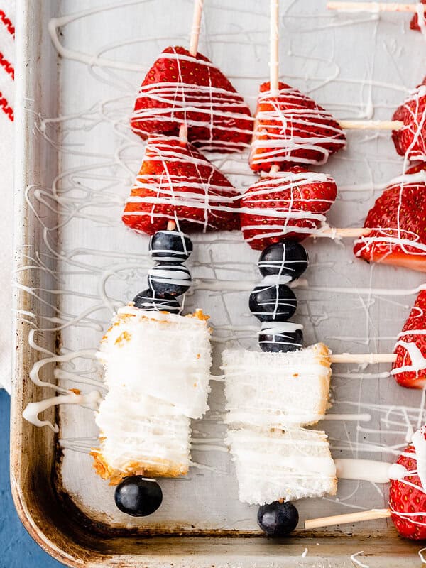 overhead dessert kabobs on baking sheet made with blueberries, strawberries, and angel food cake, drizzled with white chocolate