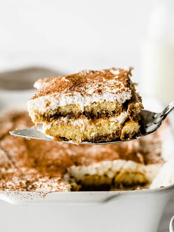 Piece of Tiramisu dessert on metal serving spoon over white casserole dish