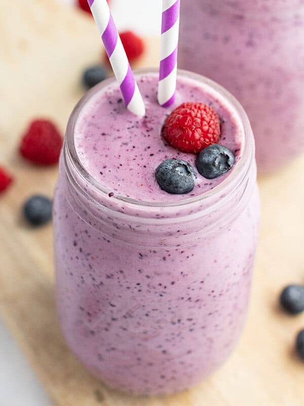 blueberry smoothie in glass garnished with blueberries, raspberries, and purple white striped straw