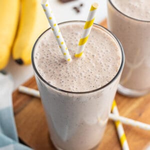 banana smoothie in tall glass with yellow and white striped straw and bananas in the background