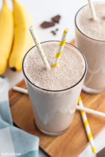 banana smoothie in tall glass with yellow and white striped straw and bananas in the background