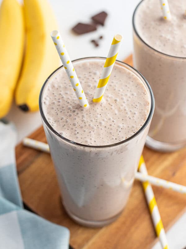 banana smoothie in tall glass with yellow and white striped straw and bananas in the background