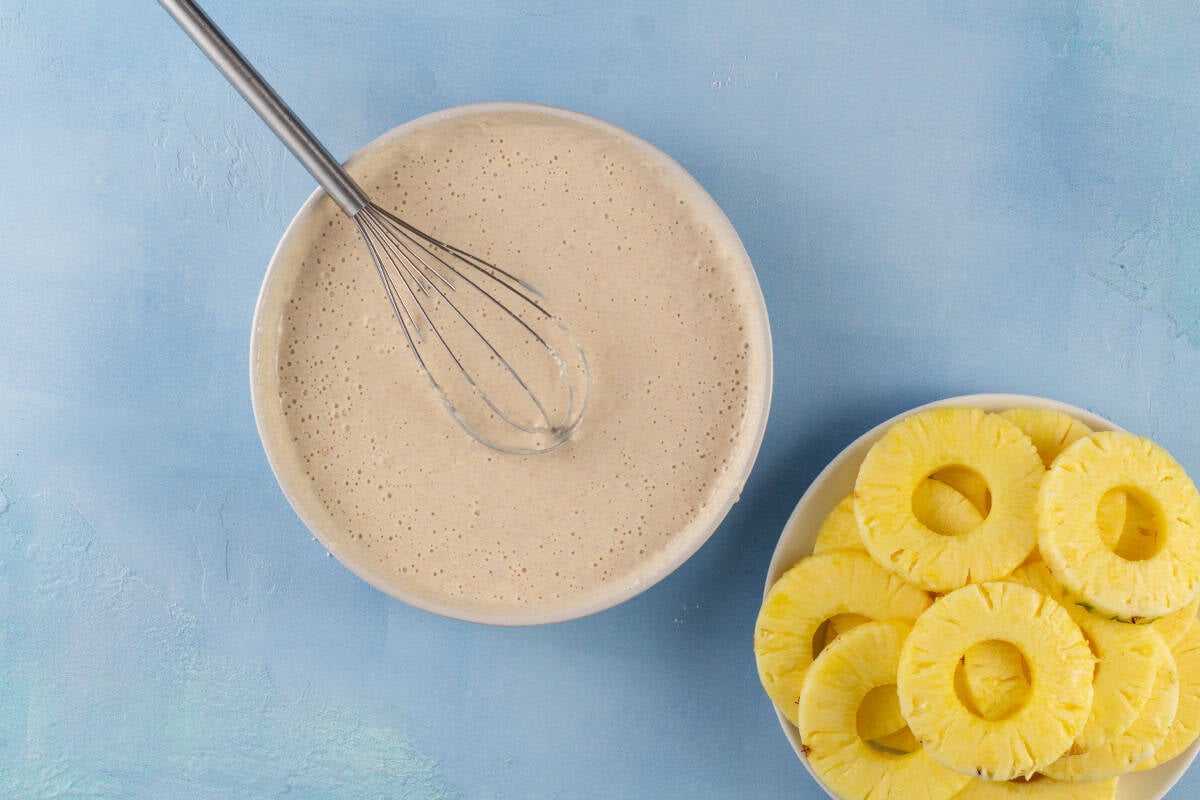 The batter for pineapple fritter rings in a white bowl with a whisk and a white bowl of pineapple rings.