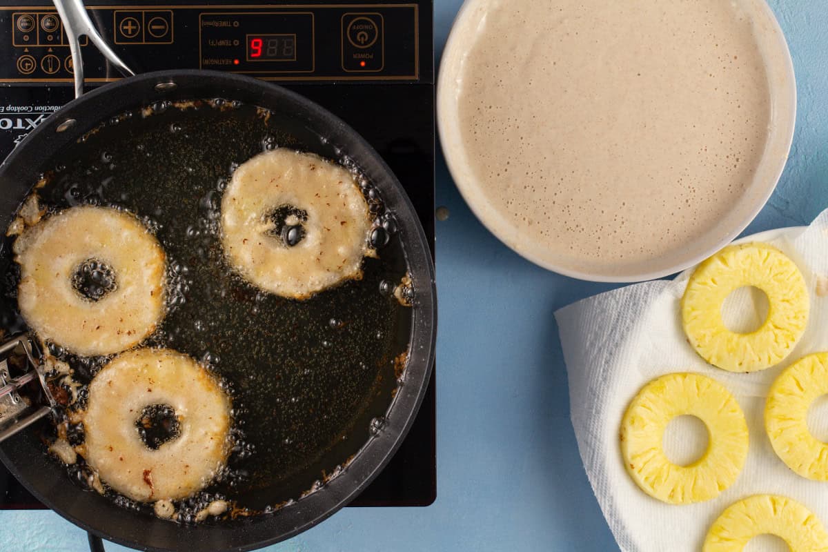 Pineapple fritters in a pan frying, a bowl of pineapple fritter batte,r and a plate of pineapple rings on a paper towel.