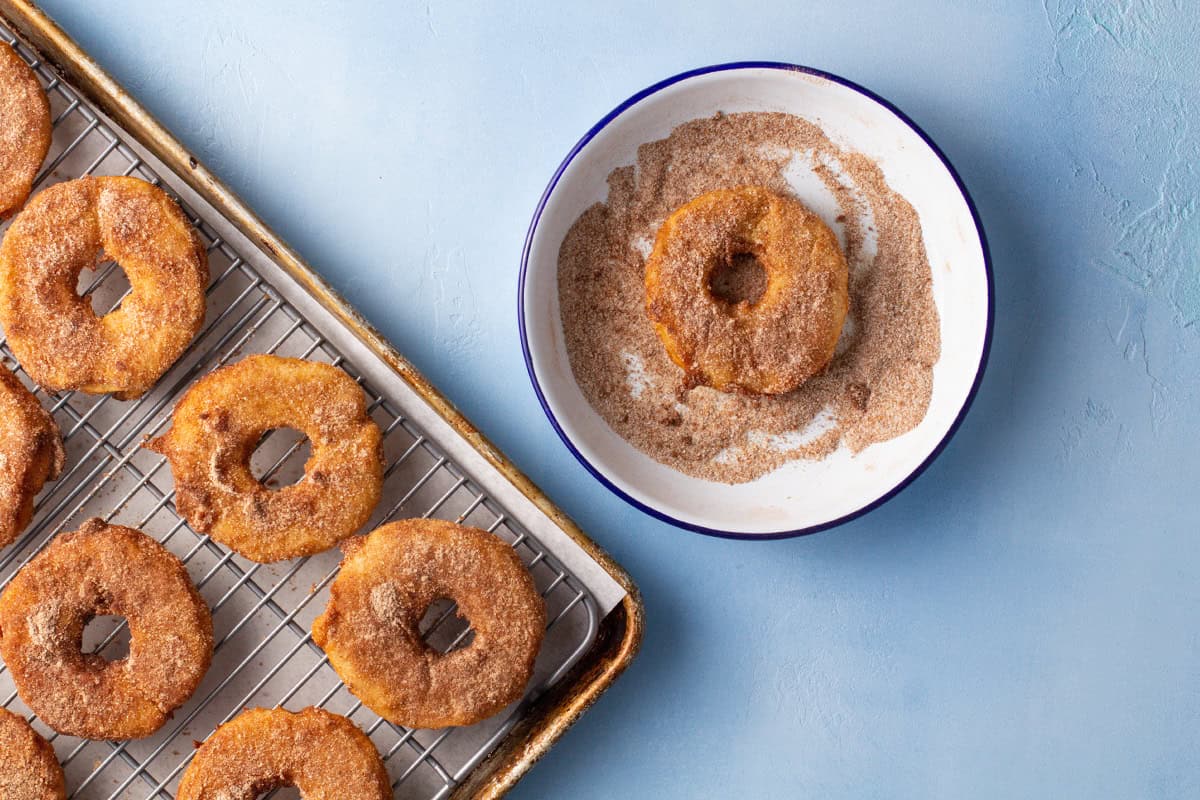 Pineapple fritter rings on a cooling rack sitting over a parchment-lined baking sheet, and a pineapple fritter in a bowl of cinnamon sugar.