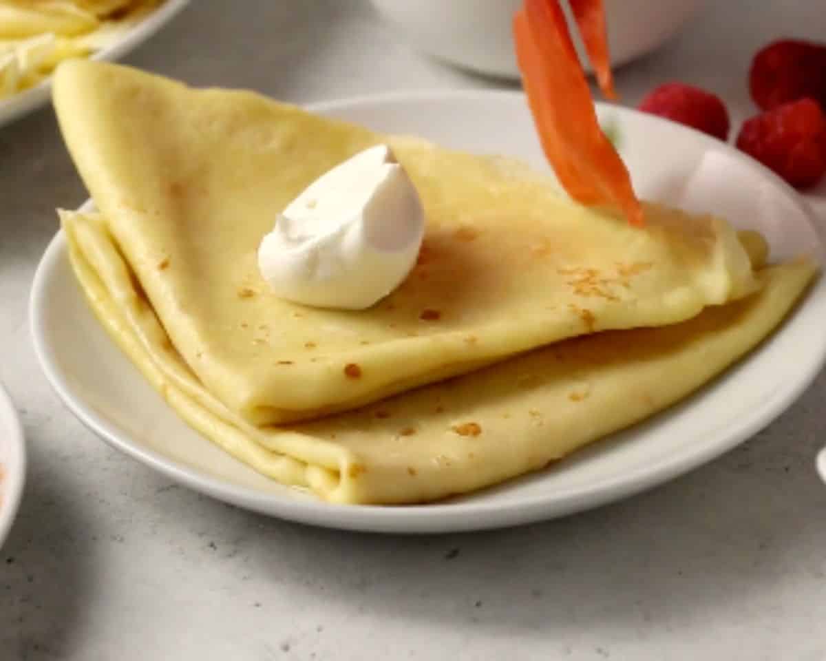 Folded blini on a white plate topped with sour cream and fresh raspberries