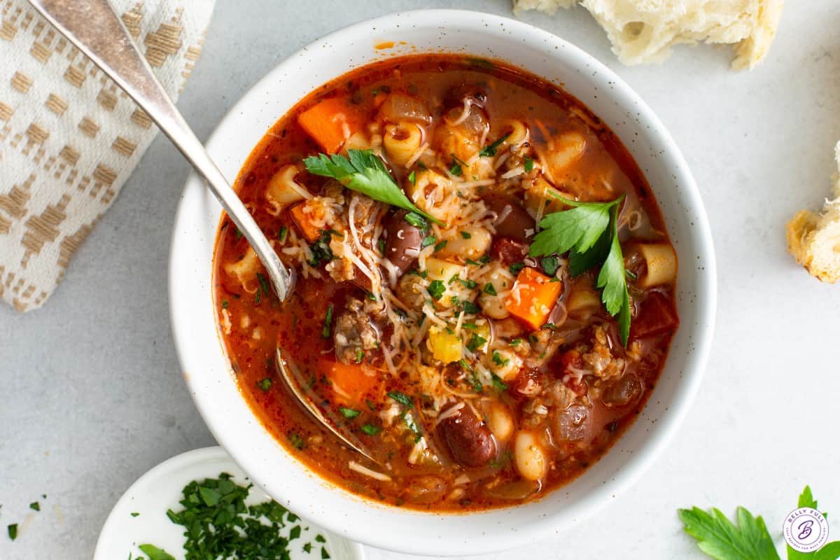 Pasta fagioli in a white bowl with a spoon, garnished with Parmesan cheese and parsley.