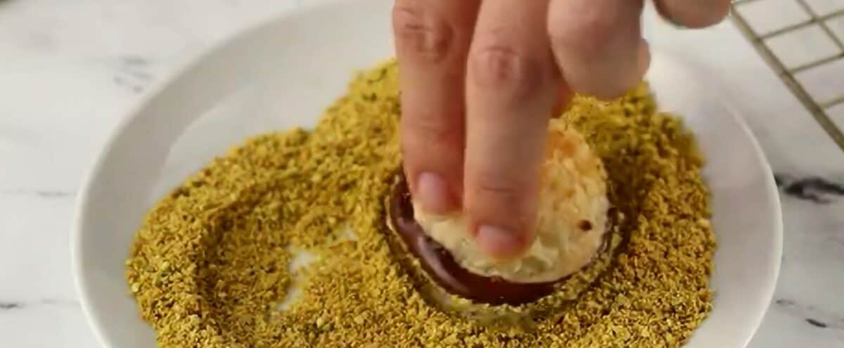 Dipping a macaroon with chocolate on the bottom into ground pistachios. 