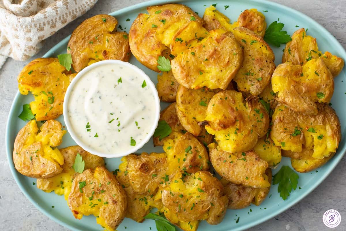 Smashed baby new potatoes on a blue serving platter with a bowl of tzatziki.