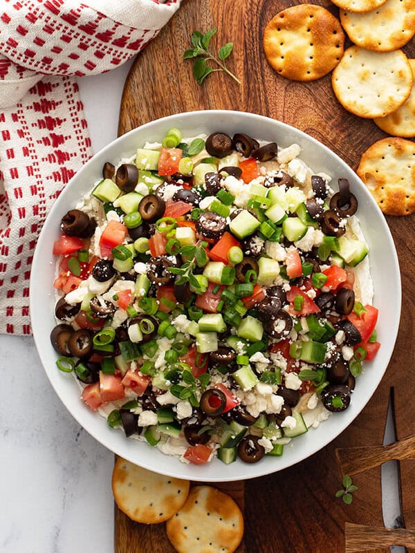 layered dip with cream cheese, hummus, olives, cucumber, and tomato