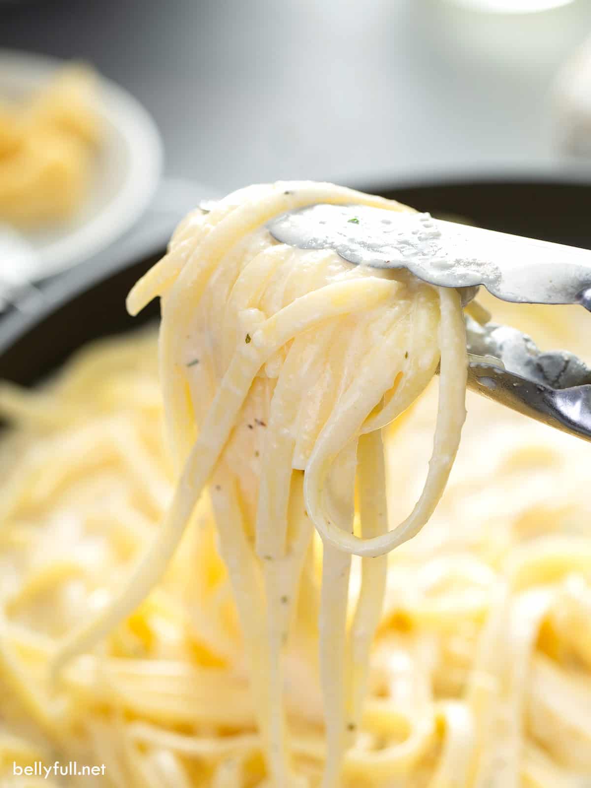 tongs holding cooked fettuccine pasta coated in creamy white sauce