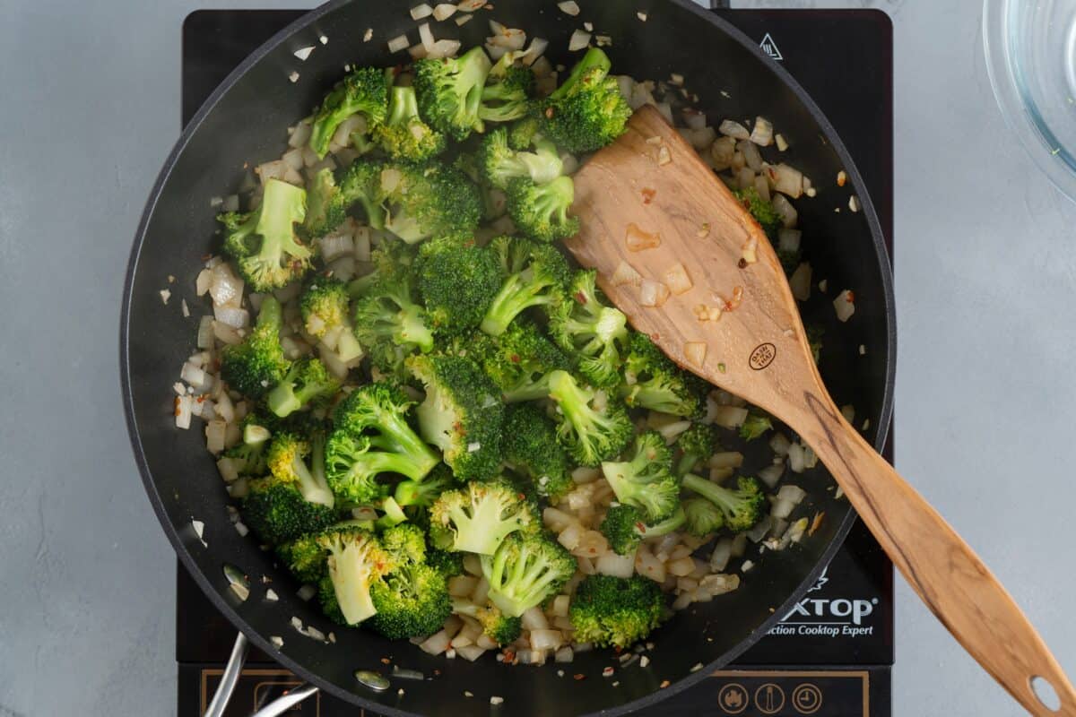 Sauteeing onions and broccoli in a skillet with a wooden spoon.