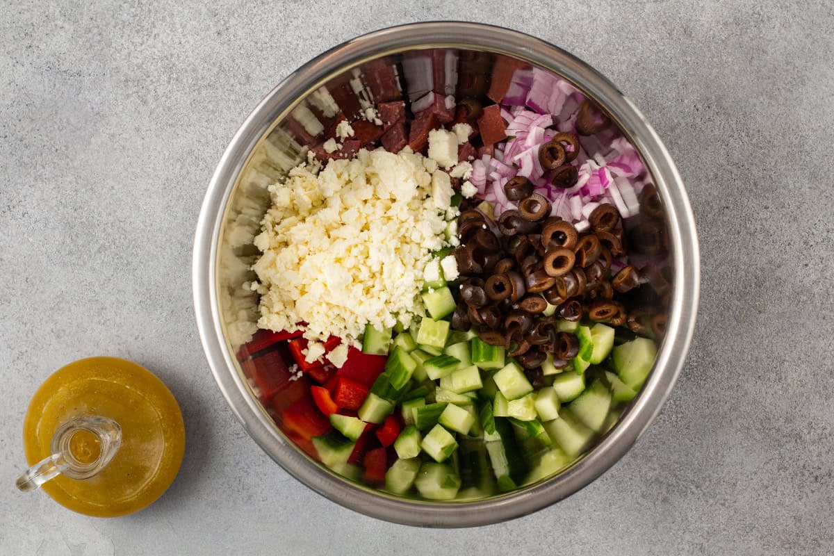 Ingredients to make a Greek pasta salad in a bowl with the dressing in a bottle on the side.