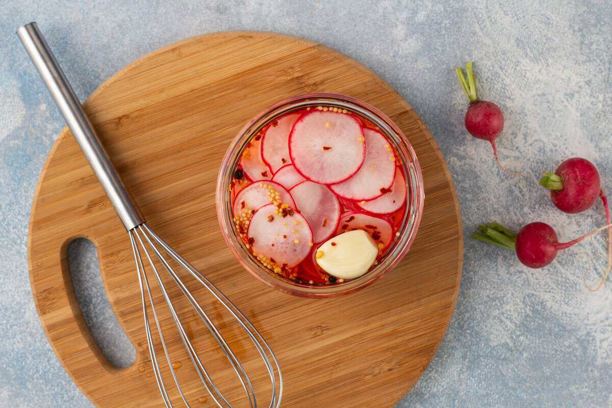 Sliced radishes in a jar with pickling spices, and brine next to a whisk, all sitting on a wooden cutting board.
