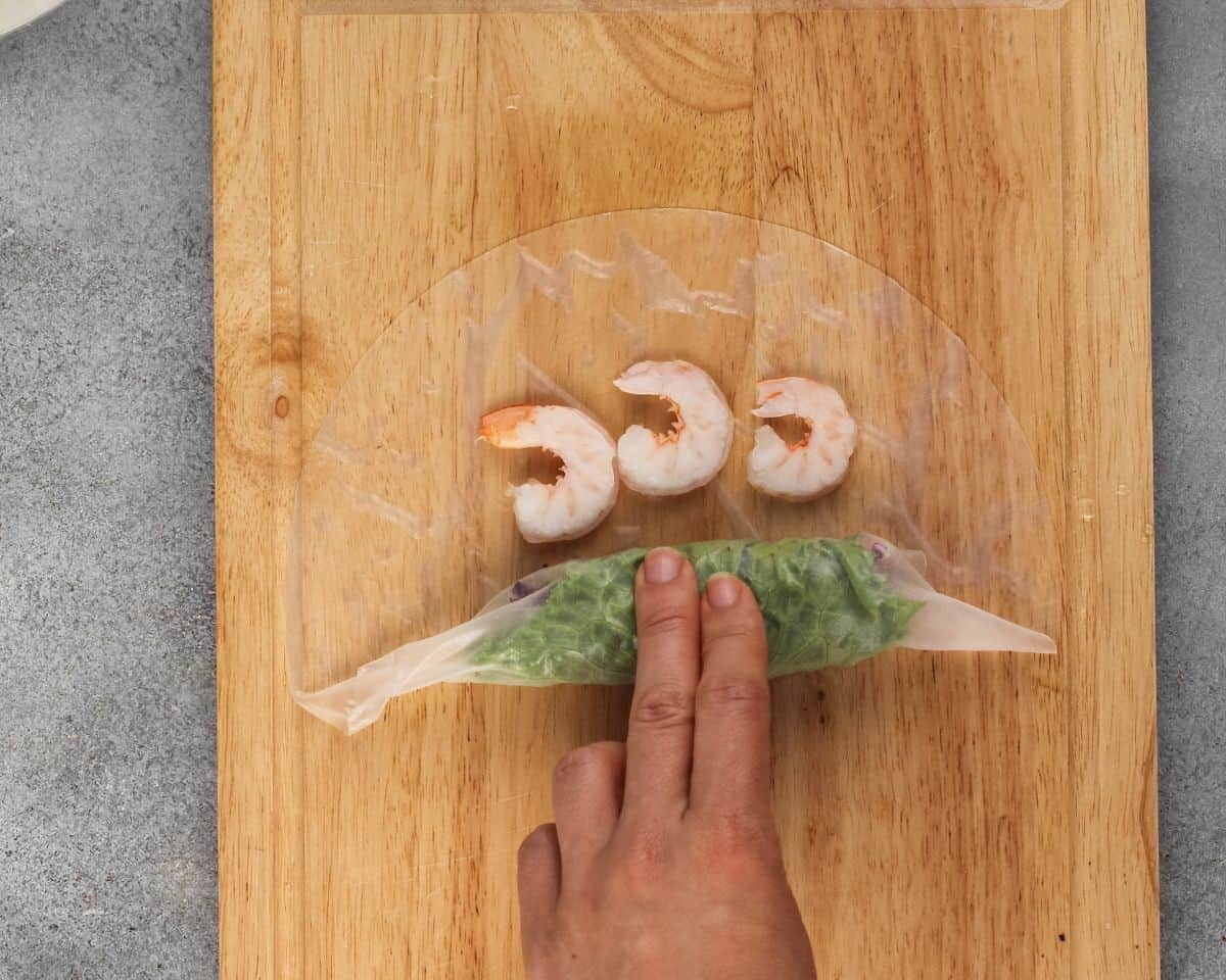Rolling the rice paper with 3 shrimp on it on a wooden cutting board.
