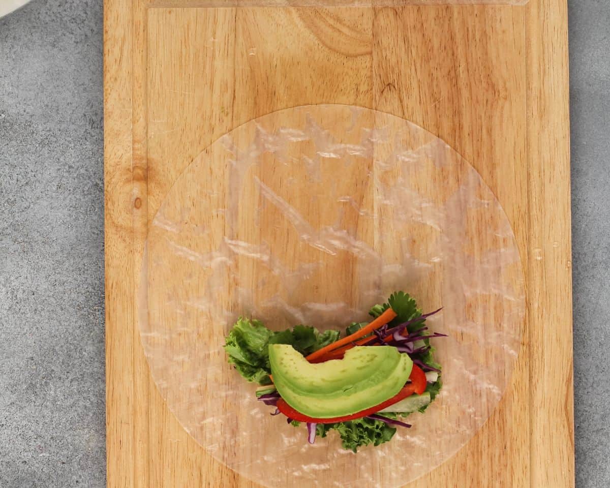 Veggies layered on top of the lettuce on the rice paper on a wooden cutting board.
