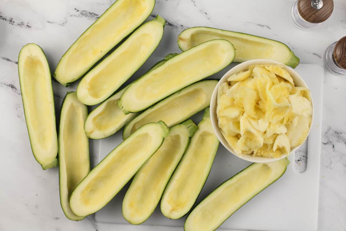 Hollowed out zucchini and a bowl of the zucchini filling on a white cutting board.