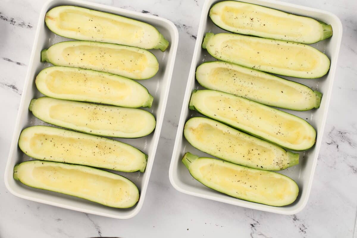 Hollowed out zucchini sprinkled with salt and pepper in 2 white baking dishes.