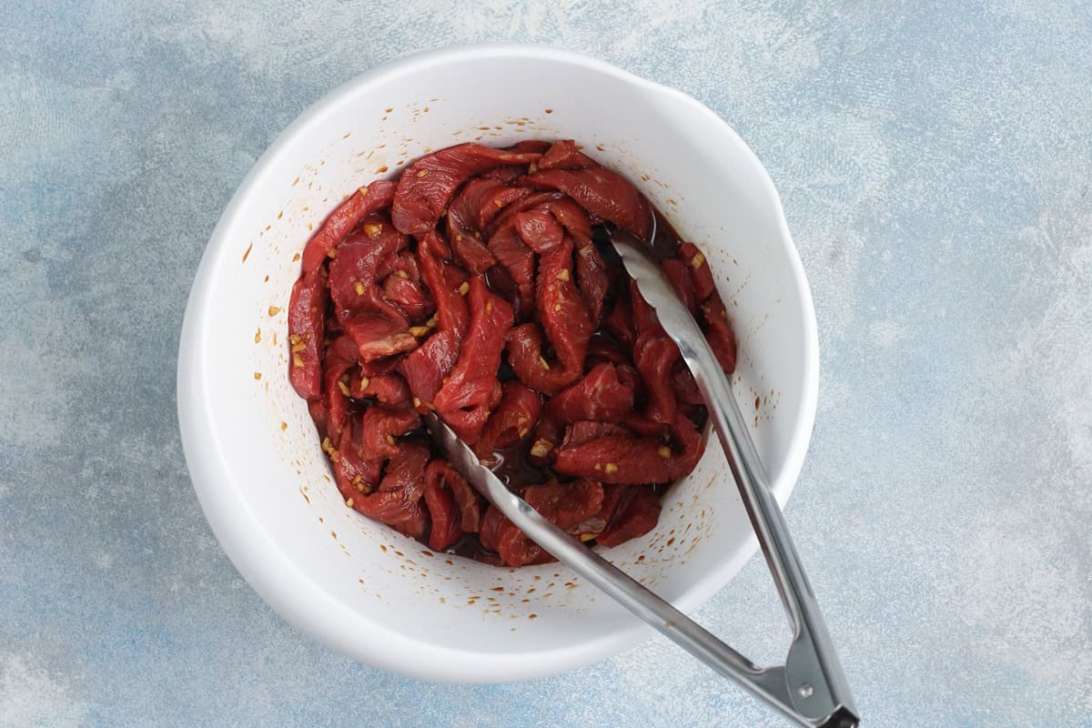 Beef marinating in a white bowl with tongs.