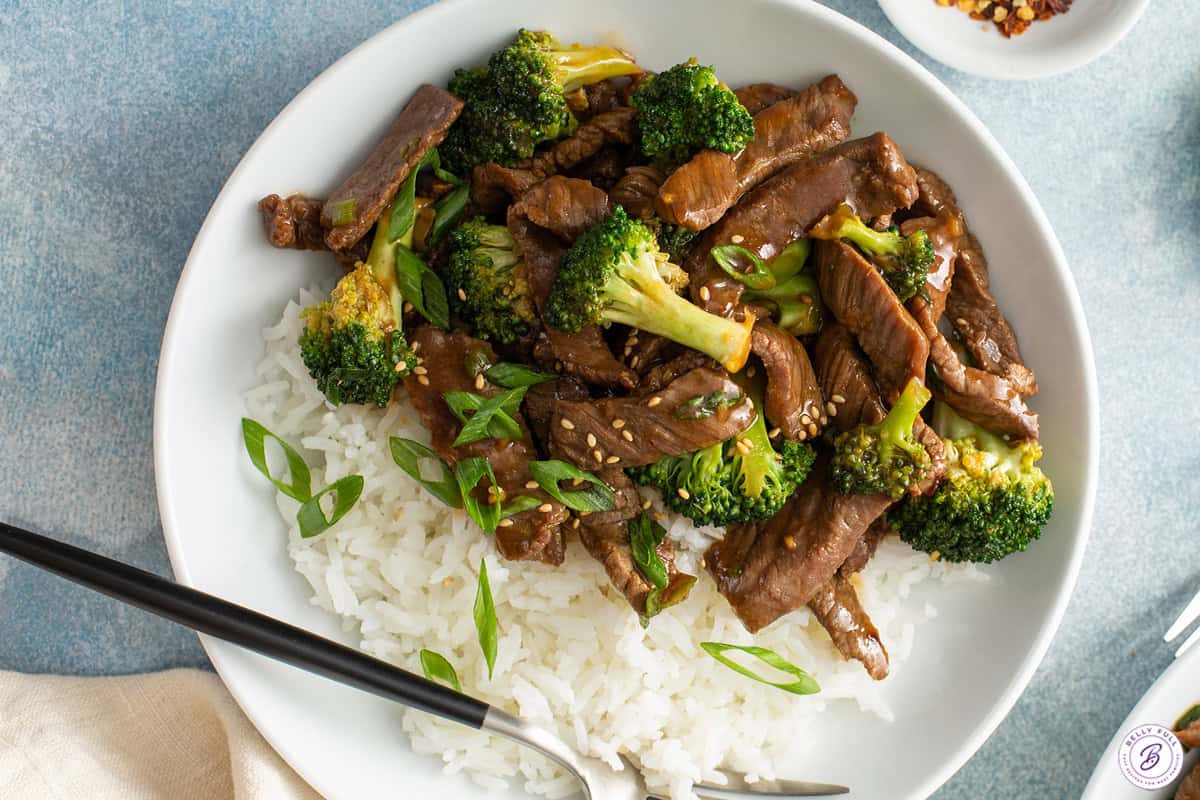 Beef and broccoli stir fry on a white plate with white rice and a fork.