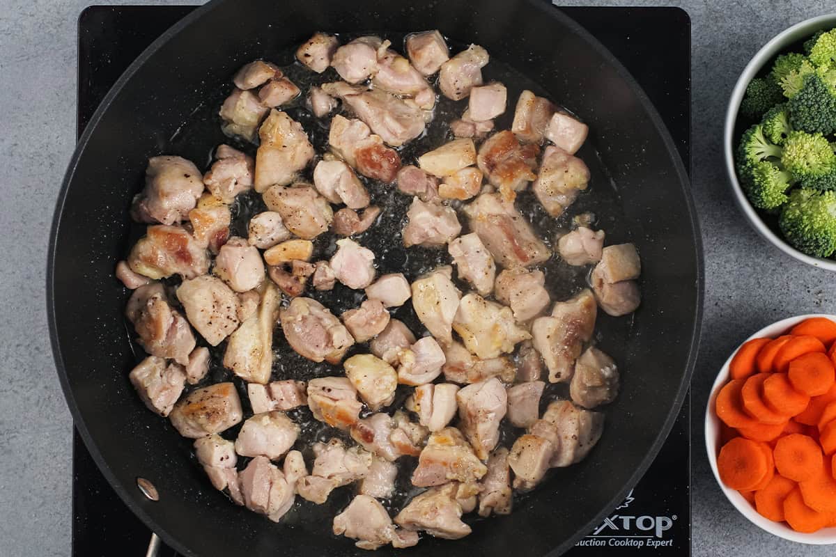 Cooking cubes of chicken in a skillet with a bowl of carrots and a bowl of broccoli next to it.