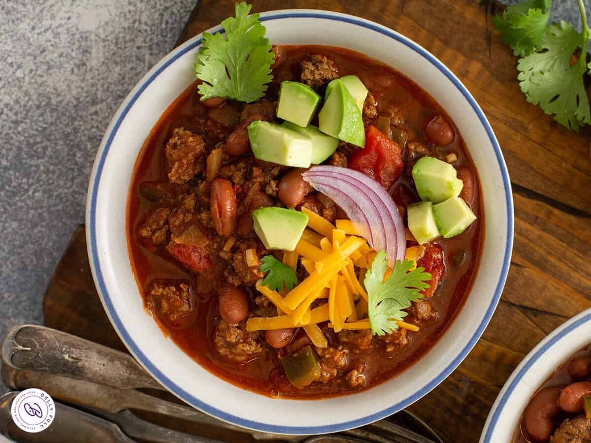 Bowl of chili garnished with cheese, avocado, red onion, and cilantro.
