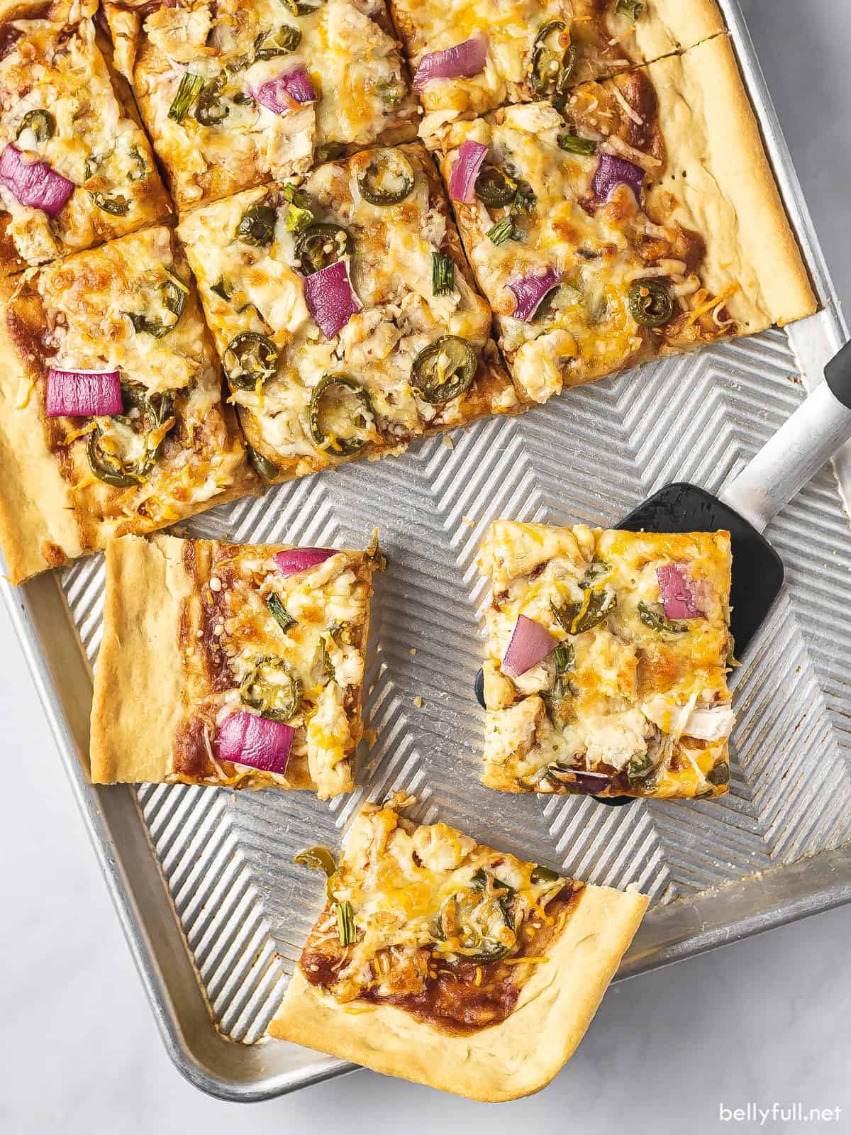 Overhead view of a sheet pan pizza with BBQ chicken and several slices missing, one being served