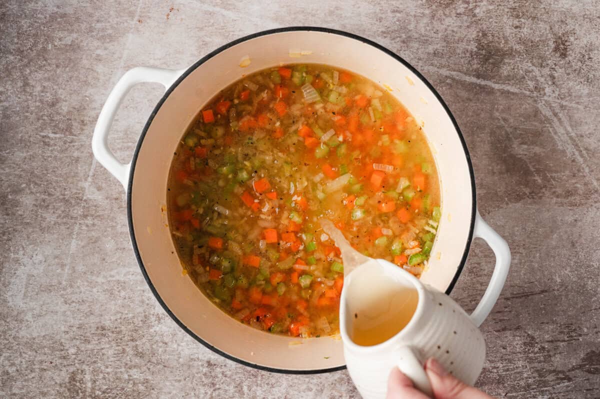 Chicken and beef broth being poured into sautรฉed vegetables in Dutch oven