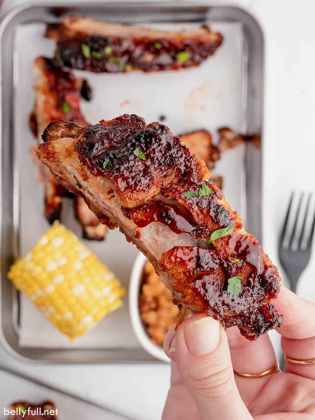 A hand holding a baby back rib over a baking sheet with other ribs