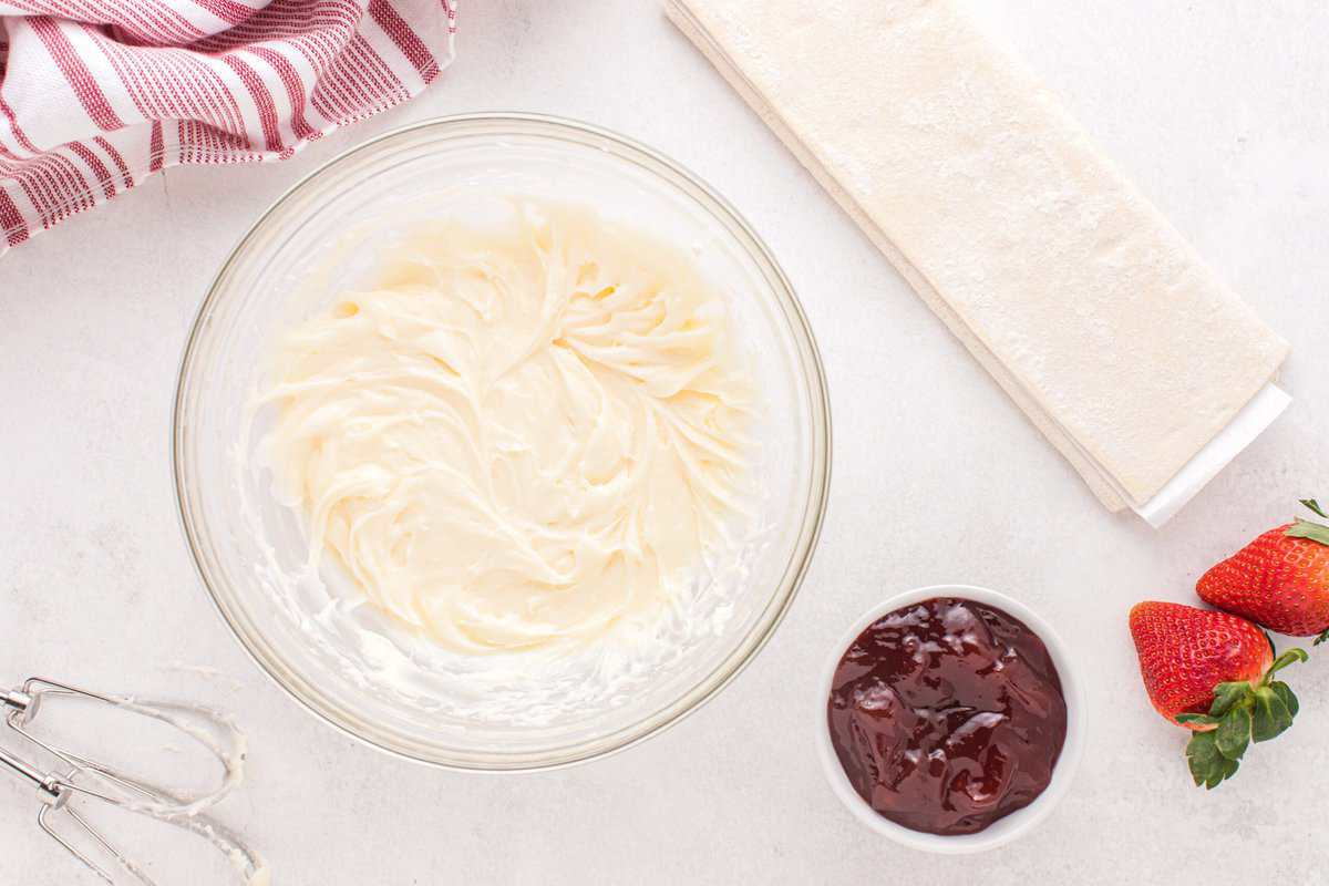 Cream cheese mixture in a glass bowl with a small bowl of strawberry jam, strawberries, and beaters from a mixer.