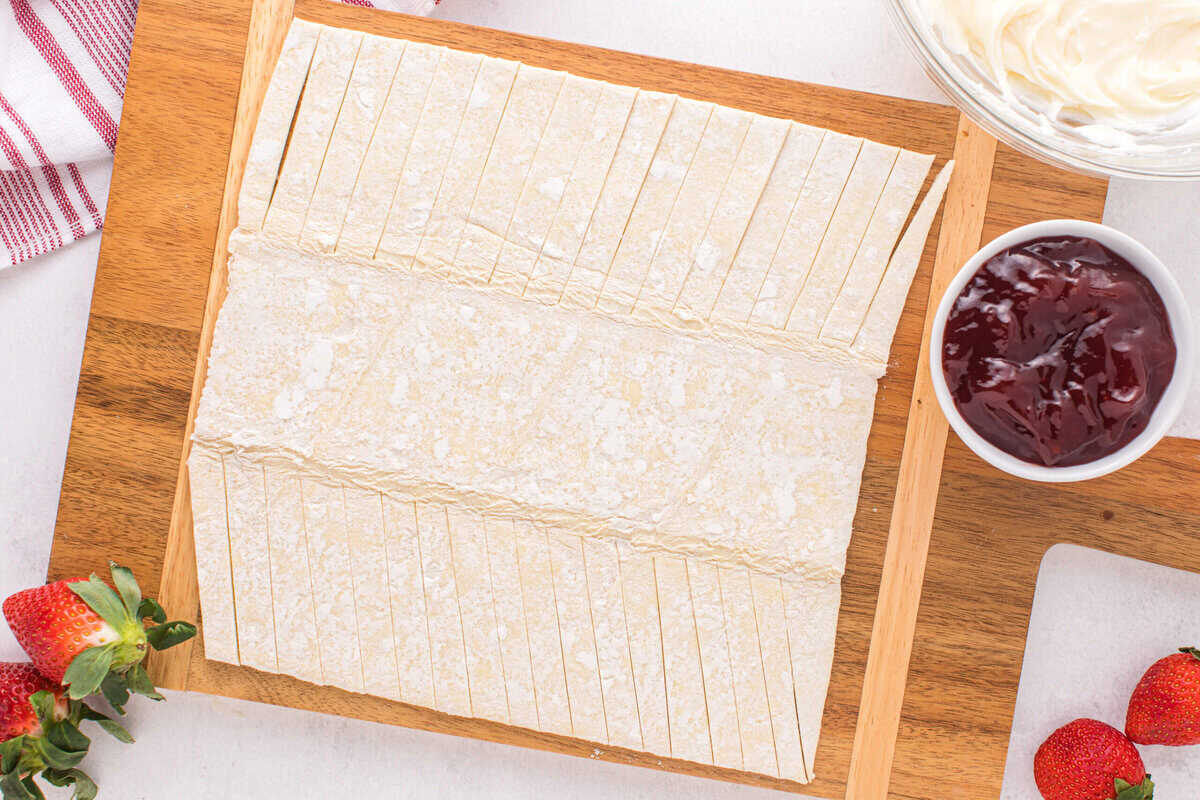 Puff pastry sheet with strips cut on a wooden cutting board, a white bowl of strawberry jam, and strawberries.