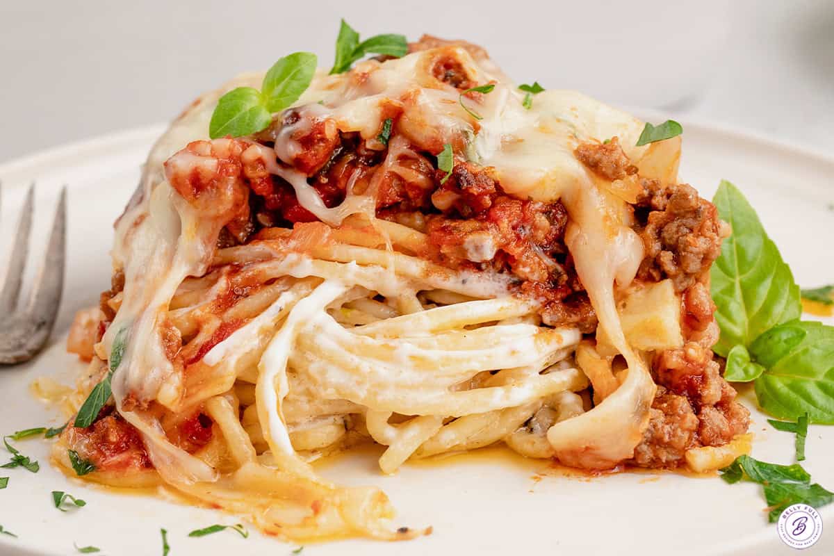 Made with spaghetti, marinara meat sauce, and homemade alfredo sauce, this Million Dollar Spaghetti Alfredo is an easy weeknight dinner. Portion of million dollar spaghetti on a white plate with a fork and basil leaves.