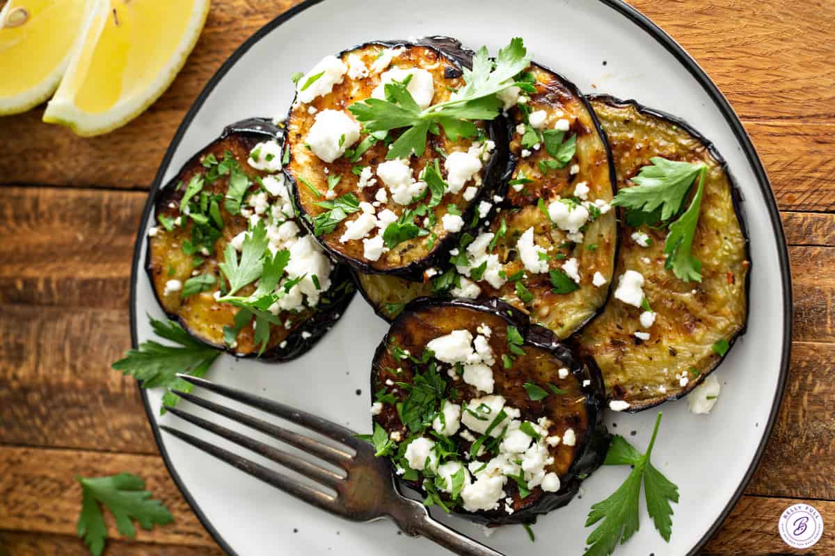 Grilled eggplant topped with feta and parsley on a white plate with a fork.