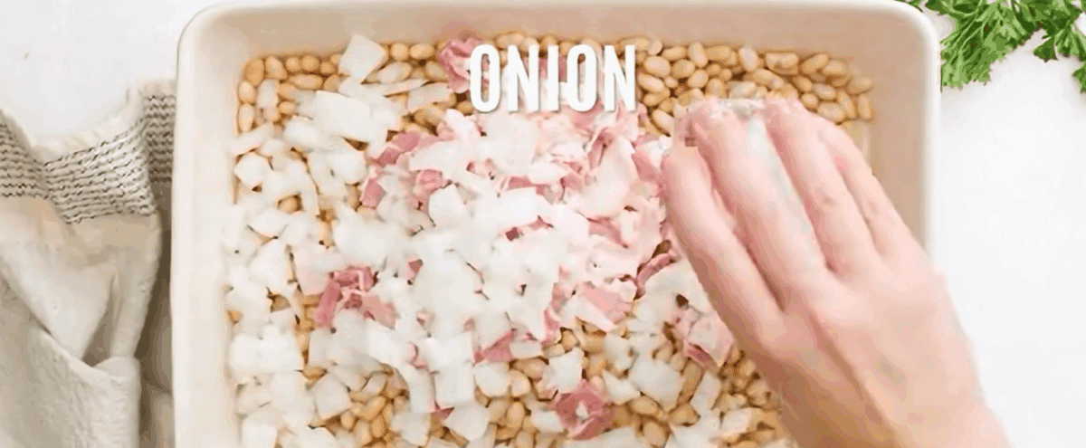 Adding beans, bacon and onion to a casserole dish. 