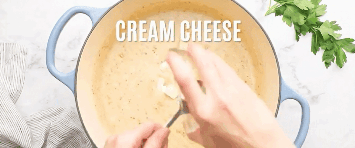 Adding cheeses and seasoning to the roux in a pot with blue handles. 