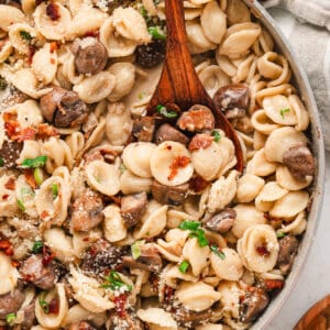 A wooden spoon stirring a pan of bacon and mushroom pasta