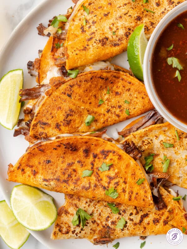 overhead overlapping Birria Tacos on plate with sliced lime wedges