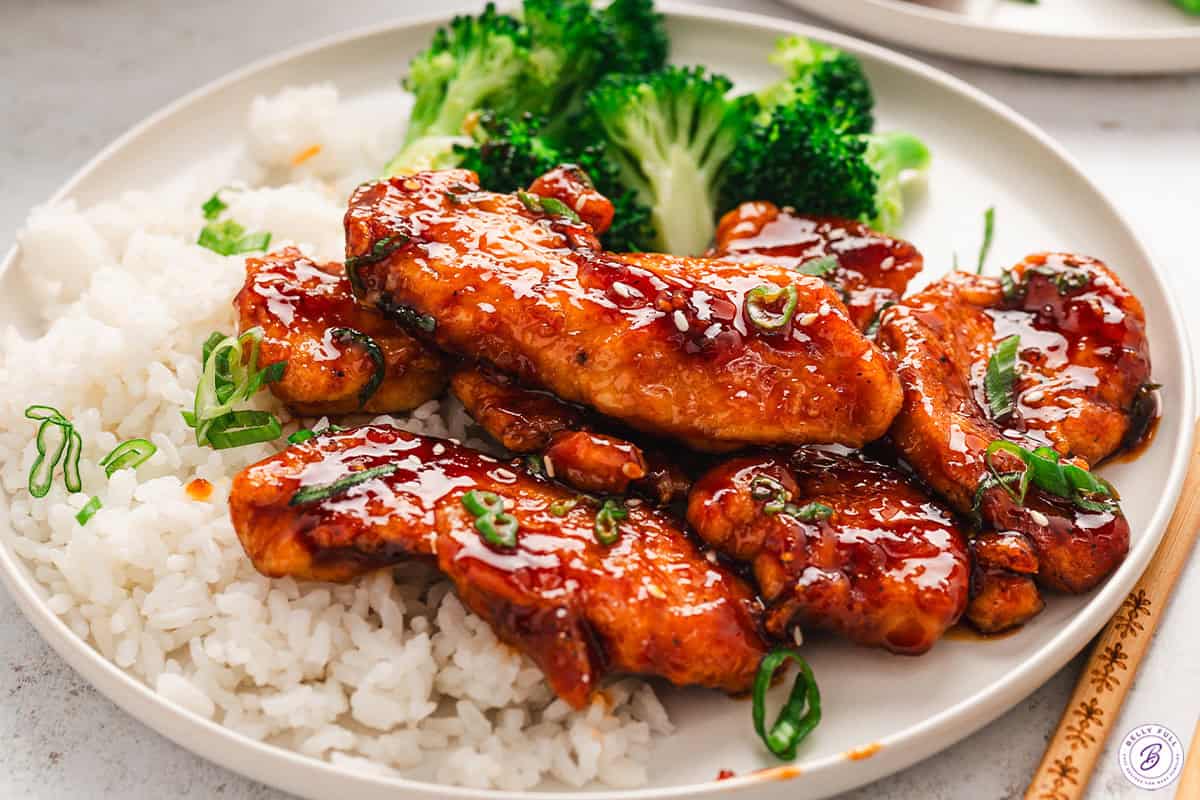 Sweet and spicy sticky chicken on a white plate with white rice and broccoli. 
