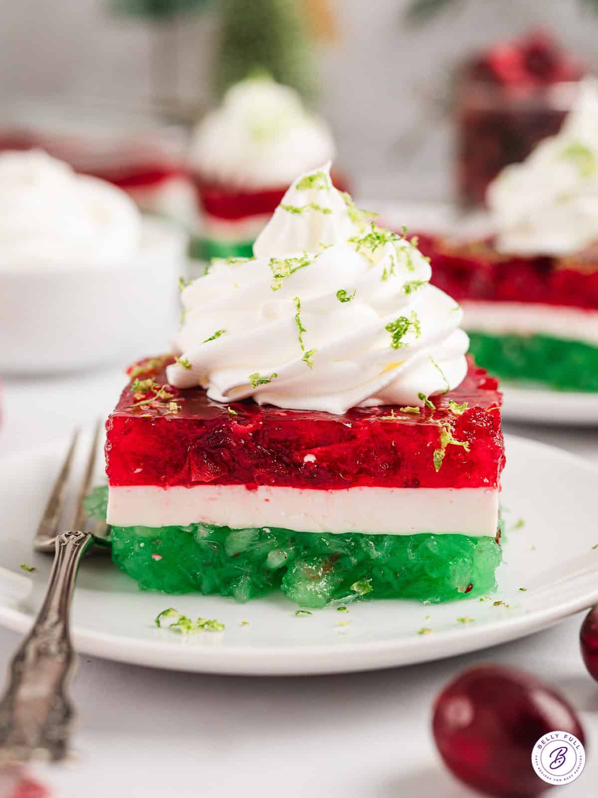 A slice of layered Christmas jello topped with whipped cream on a plate