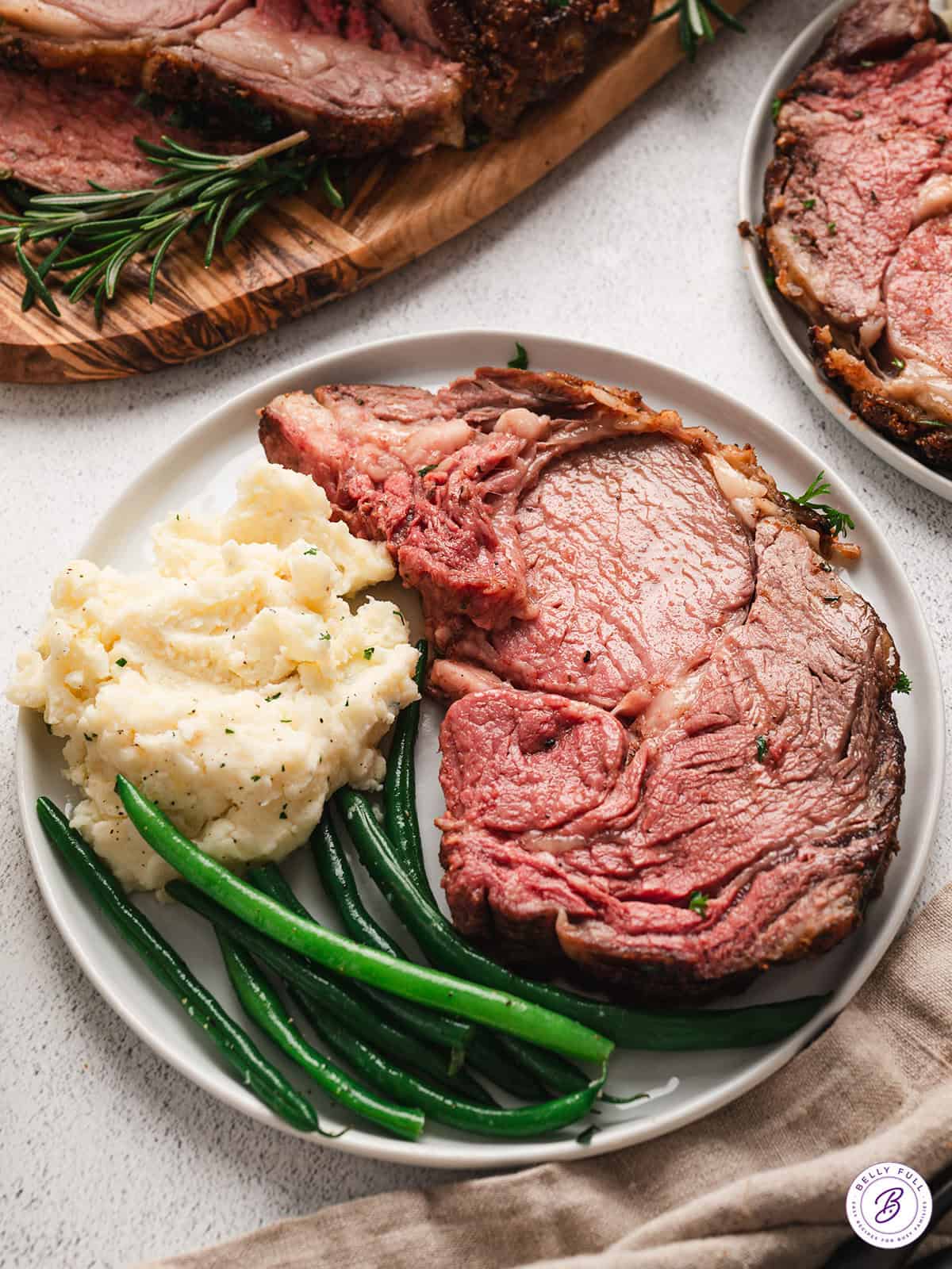 A slice of prime rib roast on a plate with mashed potatoes and green beans.